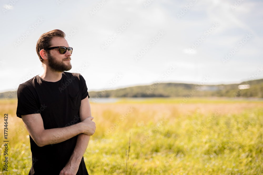 Handsome man wearing a black tshirt and jeans with dark sunglasses, standing alone on an empty field on a sunny summer day. Handsome man wearing a black tshirt and jeans with dark sunglasses, standing alone on an empty field on a sunny summer day.