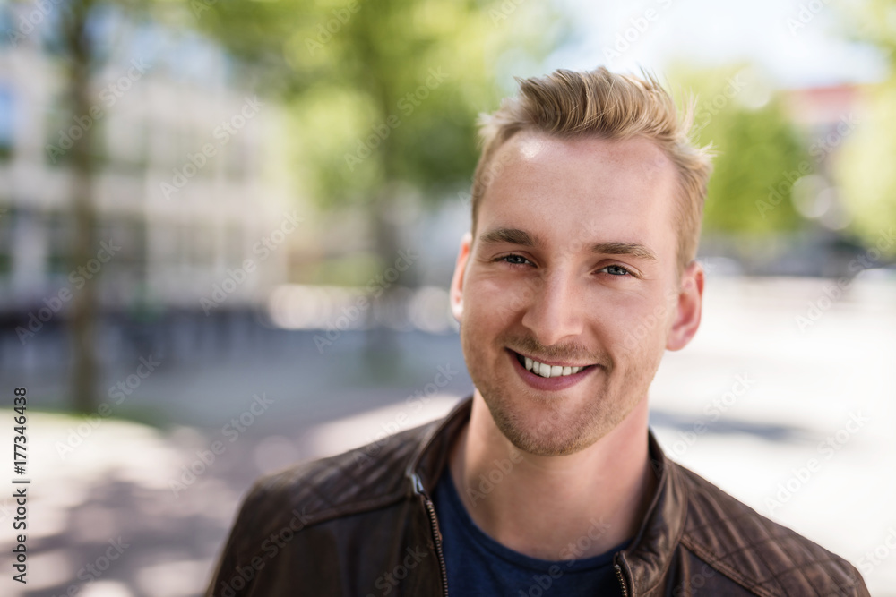 Trendy blonde man standing outdoors in a city, with trees around him wearing a brown leather jacket smiling towards camera. Trendy blonde man standing outdoors in a city, with trees around him wearing a brown leather jacket smiling towards camera.