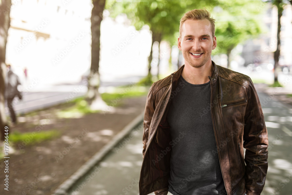 Trendy blonde man standing outdoors in a city, with trees around him wearing a brown leather jacket smiling towards camera. Trendy blonde man standing outdoors in a city, with trees around him wearing a brown leather jacket smiling towards camera.