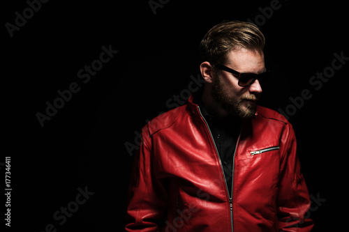 Cool man in a red leather jacket with sunglasses standing against a black background.