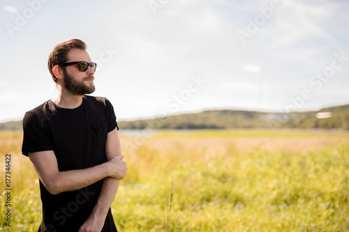 Handsome man wearing a black tshirt and jeans with dark sunglasses, standing alone on an empty field on a sunny summer day.