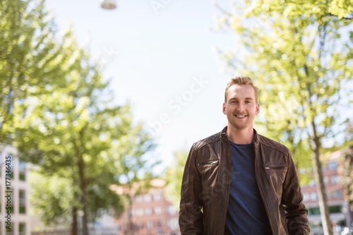 Trendy blonde man standing outdoors in a city, with trees around him wearing a brown leather jacket smiling towards camera.