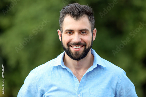 Smiling young man with dark hair and beard in blue shirt, positive expression and confident gaze, outdoor portrait