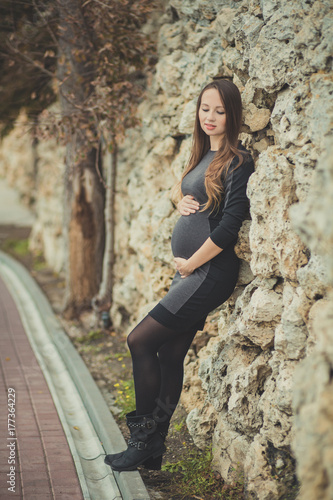 Beautiful young pregnant lady woman with brunette hair lonely dreaming closed eyes rely on stone fence wall autumn city tender holding by hands her tummy unborn baby photo