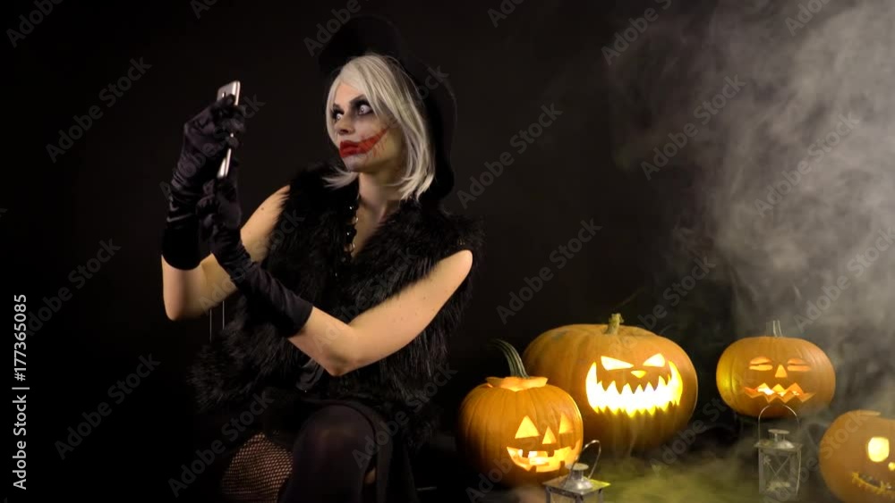 Scary beautiful girl witch celebrates halloween, makes selfie with funny glowing burning pumpkins in smoke. Woman with gray hair conjures, casts a spell. Gates of hell in fire Scary beautiful girl witch celebrates halloween, makes selfie with funny glowing burning pumpkins in smoke. Woman with gray hair conjures, casts a spell. Gates of hell in fire