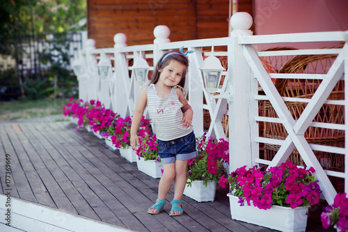 Stylish beautifull cute baby girl with brunette hair posing on wooden garden full of flowers wearing tiny jeans shirts and airy skivy underwaist and blue sandals photo