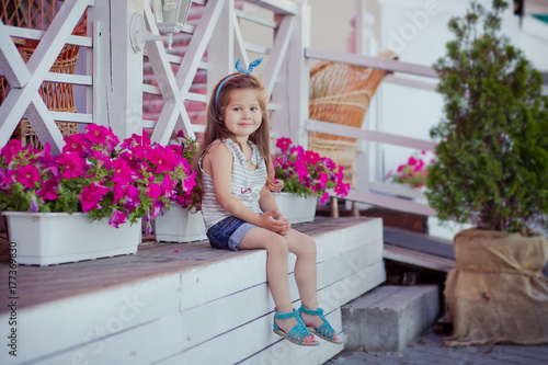 Stylish beautifull cute baby girl with brunette hair posing on wooden garden full of flowers wearing tiny jeans shirts and airy skivy underwaist and blue sandals photo