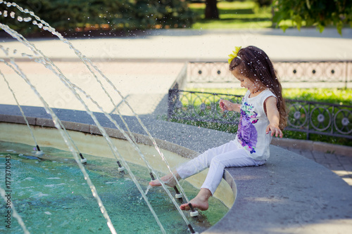 Amazing scene of little fashion baby child girl playing summer time with fountain by ner tiny barefoot legs wearing fancy clothes white colourful stuff