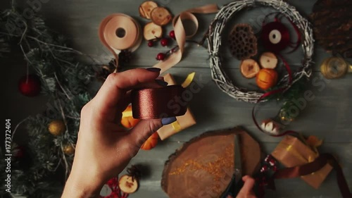 A top-down plan. Fully visible the table with the decorations. Female hands put and finalize Christmas gift wrapped in craftool paper on a wooden table. Bandaging tape and tied in a bow. photo