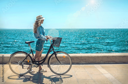Blonde woman in summer hat with her bicycle walking coastline by