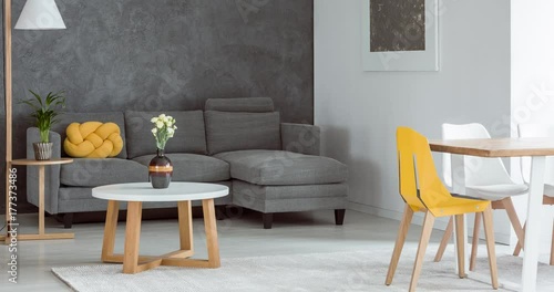 Grey corner sofa against concrete wall in living room with vase on white round table and yellow chair at dining table