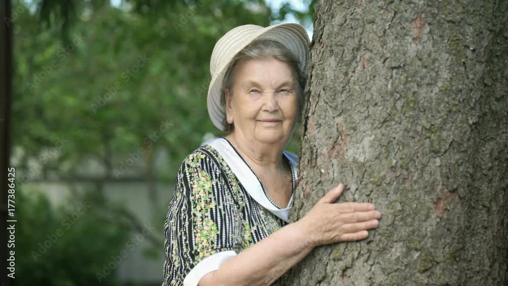 Elderly mature woman aged 80s dressed in white hat hugging tree trunk her hands smiles for camera in the park. Woman is charged energy of nature. Relaxation in a summer day Elderly mature woman aged 80s dressed in white hat hugging tree trunk her hands smiles for camera in the park. Woman is charged energy of nature. Relaxation in a summer day
