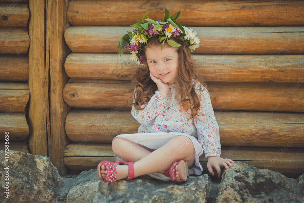 Obraz premium Cute young girl with brown eyes brunnette plait hair and pink cheek wearing white dress shirt and posing close to wooden house fence wagon looking to camera with wreath of flowers on head