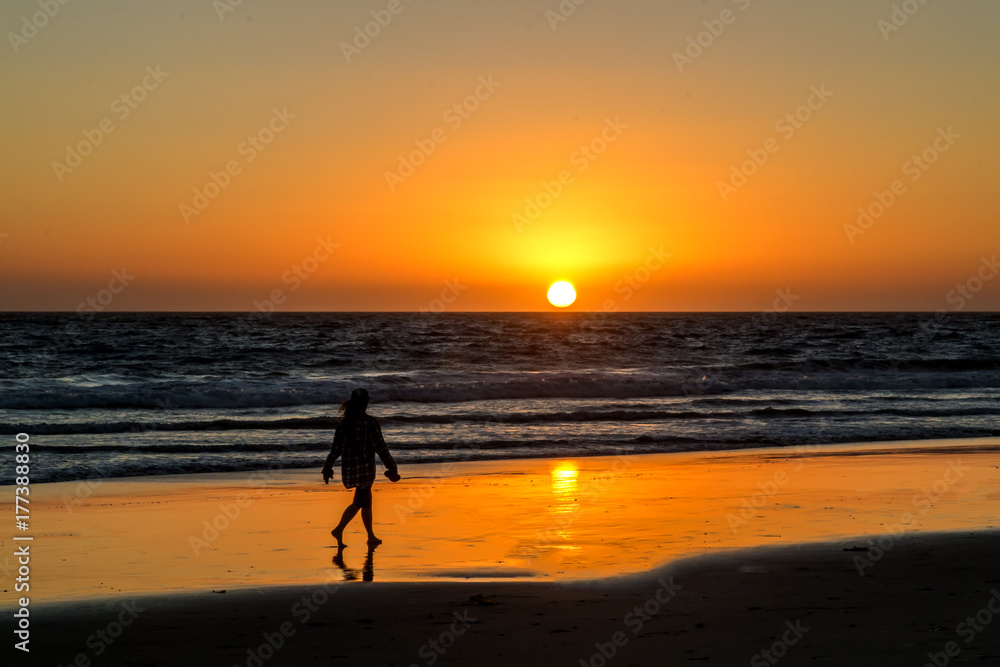 Red, Yellow and Orange Sunset Over Wave covered Ocean with Woman Walking in Wet Sand Reflecting Sun