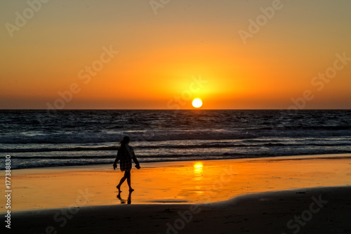 Red, Yellow and Orange Sunset Over Wave covered Ocean with Woman Walking in Wet Sand Reflecting Sun