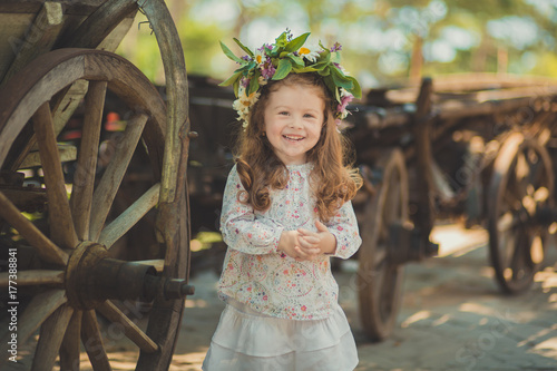 Cute young girl with brown eyes brunnette plait hair and pink cheek wearing white dress shirt and posing close to wooden house fence wagon looking to camera with wreath of flowers on head photo