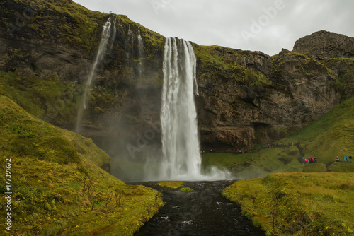 A number of steep cliffs on the right and a huge waterfall. Gloomy  severe weather  gloomy sky. Everything is shrouded in smoke from the waterfall. Nature and sights of Iceland.  