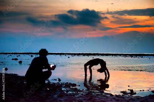 Fototapeta Naklejka Na Ścianę i Meble -  The girl shows the figures standing in the bridge, on hands with a bent back, the guy shoots video on the smartphone, against the sunset at the sea. Gili Trawangan island, Lombok, Indonesia.