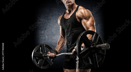 Young sexy bodybuilder with perfect body doing exercise with barbell on black background.