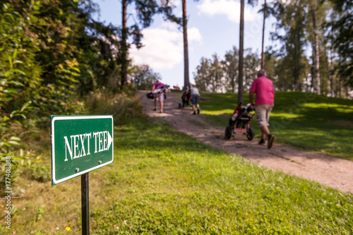 golf players walking up the hill towards the next tee with "next tee" sign