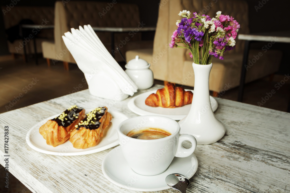 White plate with Coffee eclairs and croissant with cup of fresh black coffee on wooden table. Top view. White plate with Coffee eclairs and croissant with cup of fresh black coffee on wooden table. Top view.
