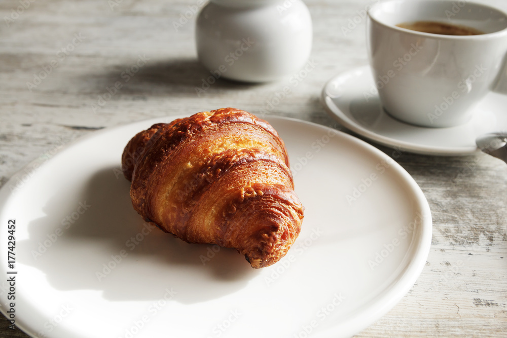 White plate with croissant with cup of fresh black coffee on wooden table. Top view.