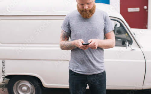 Bearded young man is texting on his cell phone with car on background photo