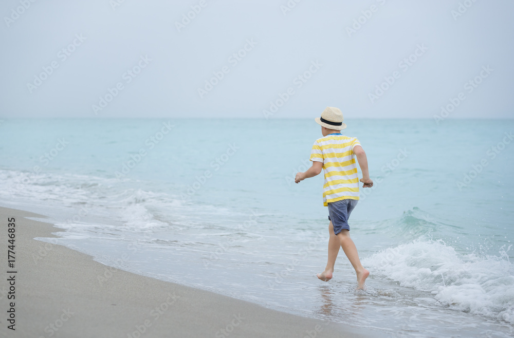 Portrait of cute kid boy running on the ocean or sea edge. Child walking on the beach and playing with waves. Lifestyle