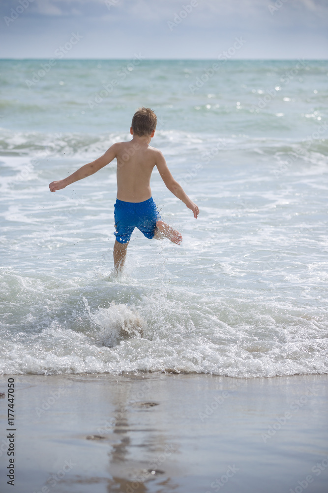 Back view on happy running kid boy rushing into ocean water on the beach. Child enjoying waves on the beach. Lifestyle Back view on happy running kid boy rushing into ocean water on the beach. Child enjoying waves on the beach. Lifestyle