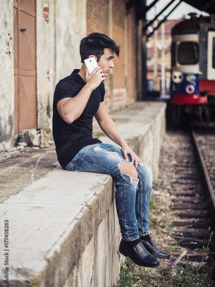 Attractive young man sitting on railroad, calling somebody on cell phone, wearing black t-shirt and jeans, looking afar Attractive young man sitting on railroad, calling somebody on cell phone, wearing black t-shirt and jeans, looking afar