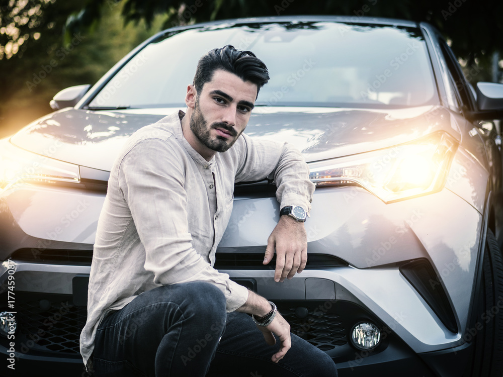 Portrait of young bearded man leaning on his new stylish polished car, outdoor