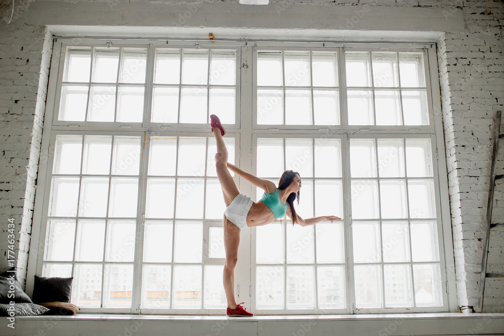 Fototapeta premium Profile portrait of beautiful young woman in aquamarine top and white shorts standing split against big window. Full length