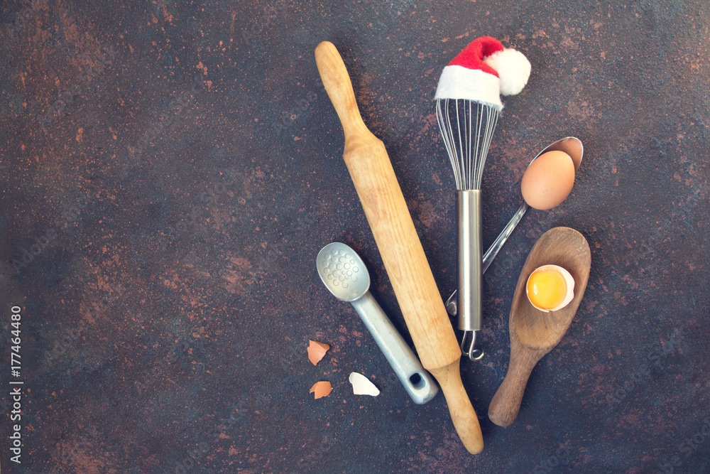 Accessories for New Year's Christmas buns whisk in a santa cap, rolling pin, spoon and eggs, holiday concept on a dark background with copy space view from above Accessories for New Year's Christmas buns whisk in a santa cap, rolling pin, spoon and eggs, holiday concept on a dark background with copy space view from above