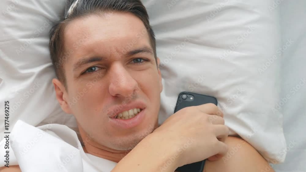 young man lies in bed, enjoys the phone and misses the smartphone on his face young man lies in bed, enjoys the phone and misses the smartphone on his face