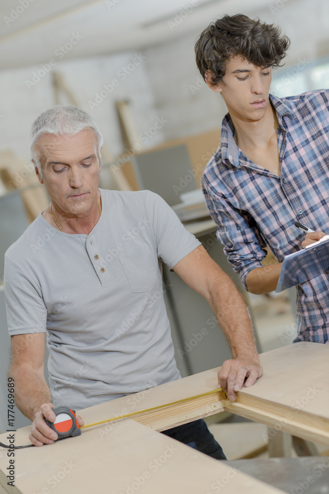 Senior carpenter measuring hole in wood, junior taking notes Senior carpenter measuring hole in wood, junior taking notes