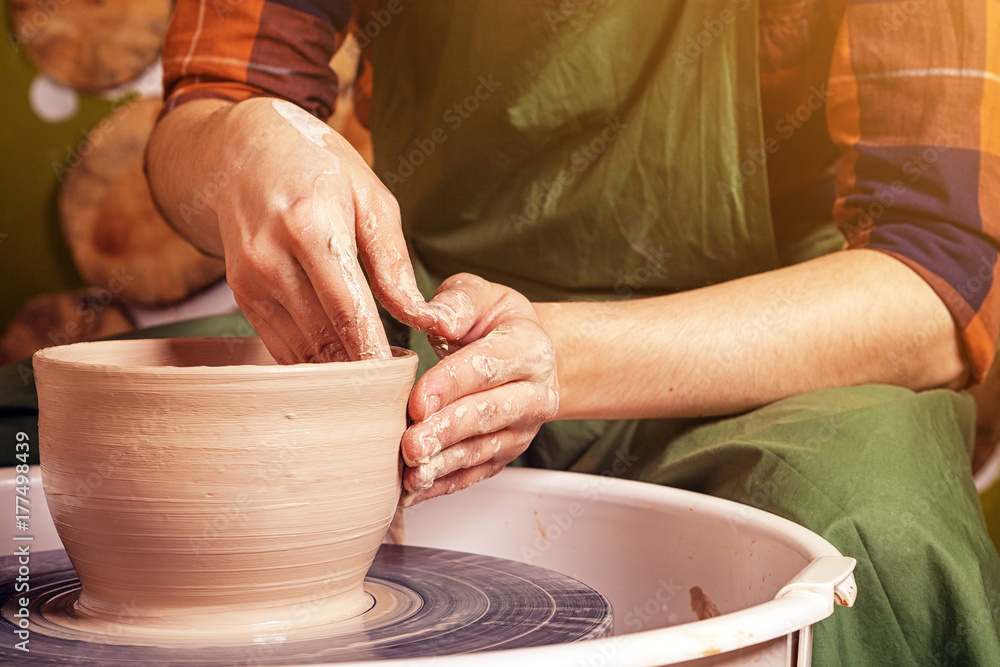 Close-up A woman potter in a plaid shirt and green apron beautifully sculpts a deep bowl of brown clay and cuts off excess clay on a potter's wheel in a beautiful workshop