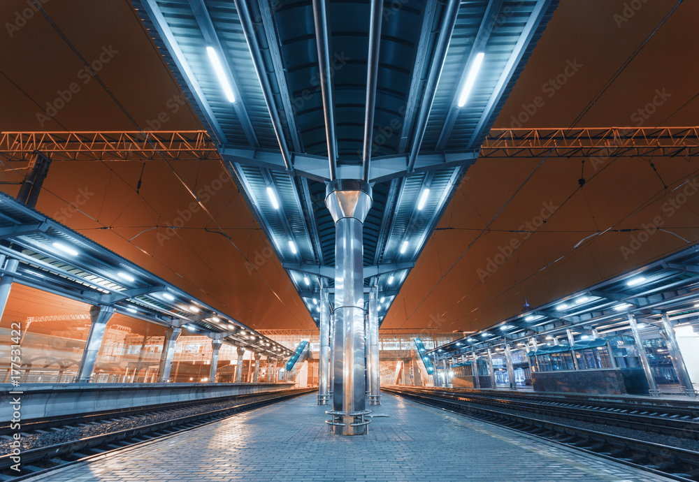 Obraz premium Modern futuristic railway station with illumination in fog at night in Europe. Amazing industrial landscape with railway platform, railroad, sky, city lights and building. Railway transportation