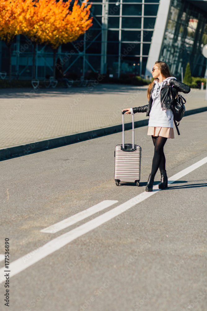 Happy cheerful traveler hipster woman standing with suitcases on the road over airport building in autumn time. Concept of travel, holidays, journey, trip.