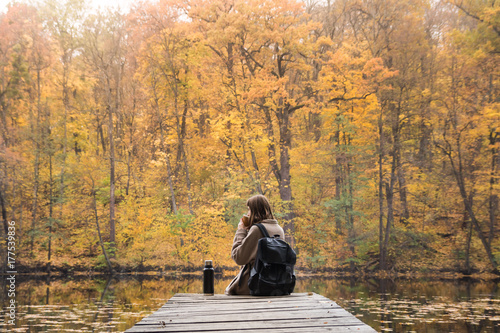 Girl sits near lake in autumn and drinks coffee. Young female person hiking at nature park rests at riverbank on gold autumn day and enjoys beautiful october scenery having a hot drink from thermos