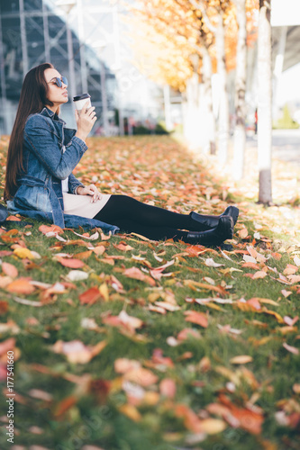 Bright picture of young fashion woman drinking coffee from disposable cup and sitting on the grass strewned with yellow and red leaves. Education, lifestyle and people concept. photo