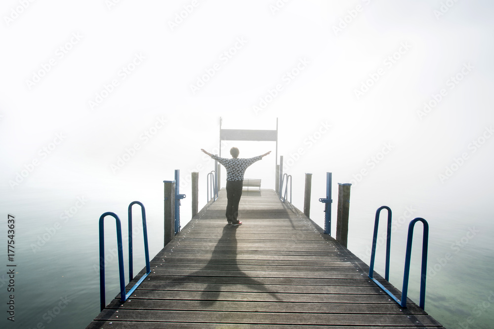 Frau steht auf Bootssteg und schaut mit erhobenen Armen auf den See im Nebel
