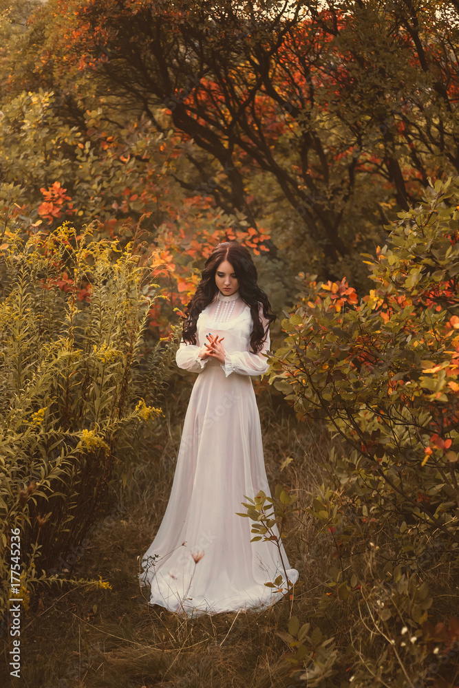 A brunette girl with luxurious, thick hair in a white vintage dress. The background is fiery autumn. Art photo