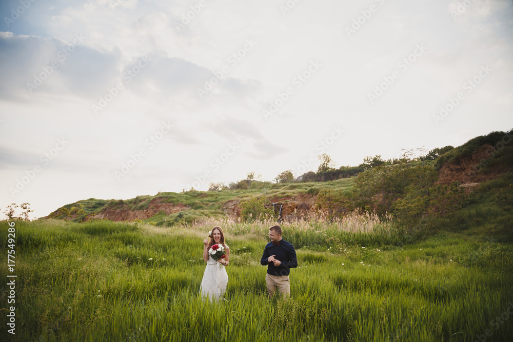 Outdoor wedding ceremony, stylish happy newlyweds are standing in green field showing fingers with wedding rings