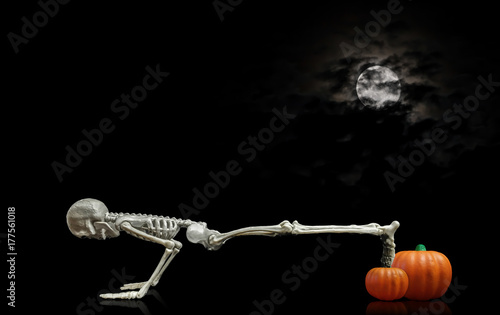 Halloween skeleton doing pushups on a pumpkin under a nice night time moon covered with dark clouds.