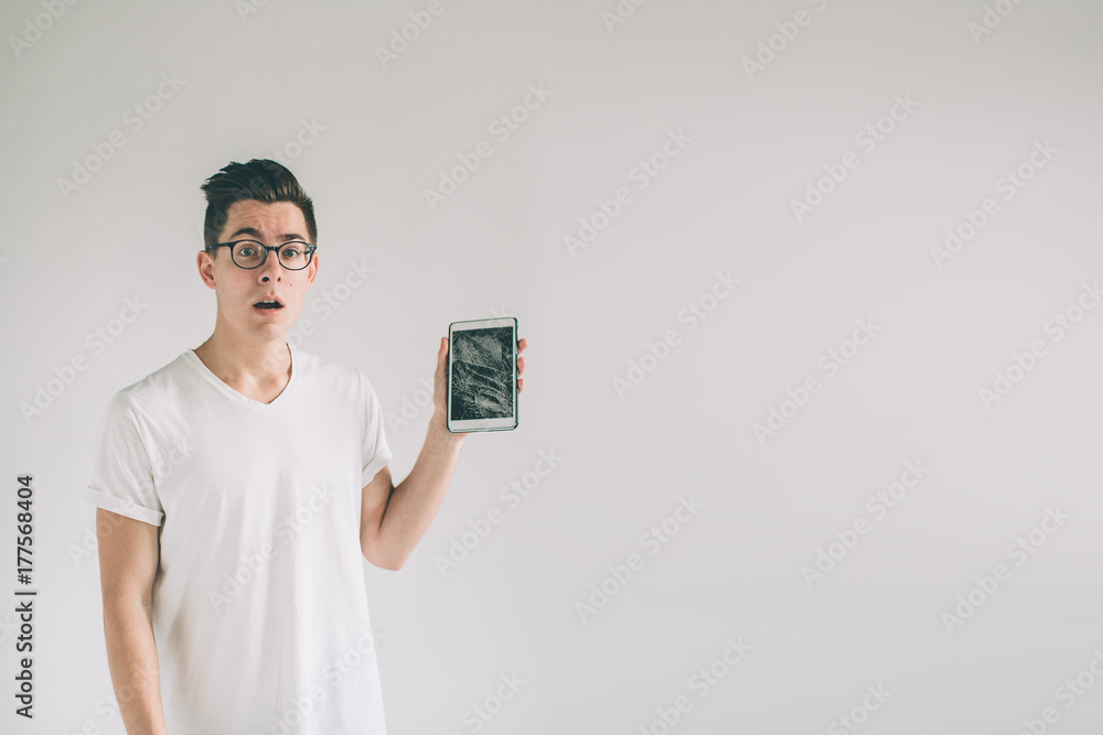 Nerd is wearing glasses. Student presenting a broken black tablet behind glass. upset man holds a out-of-use tablet or smartphone. Isolated on a light background. Broken screen Nerd is wearing glasses. Student presenting a broken black tablet behind glass. upset man holds a out-of-use tablet or smartphone. Isolated on a light background. Broken screen