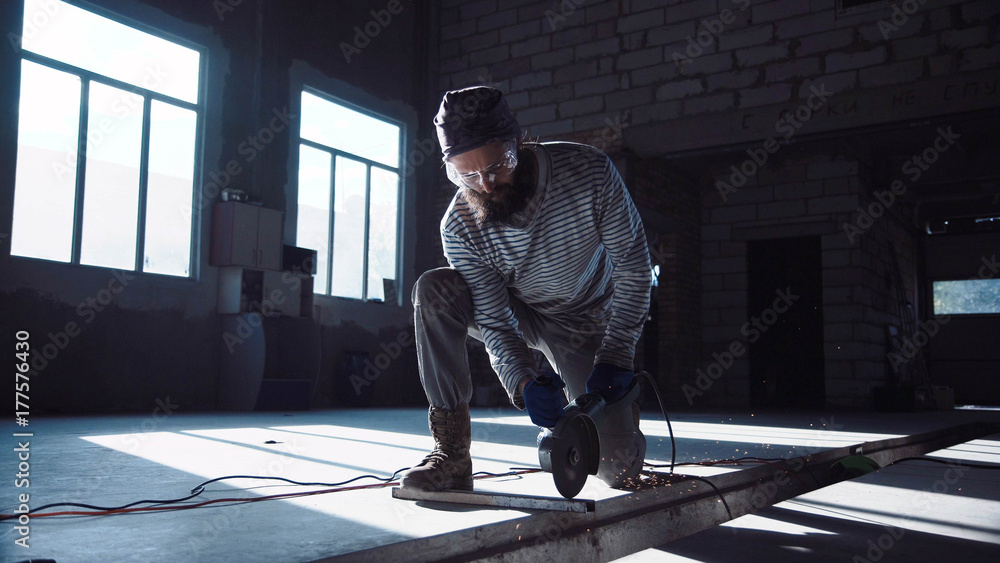 Bearded man in protective glasses working in hangar and cutting metal rod with angle grinder. Bearded man in protective glasses working in hangar and cutting metal rod with angle grinder.