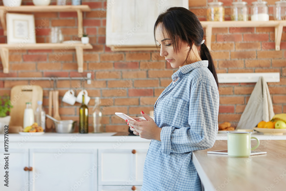 Brunette female dressed casually, has painted nails, looks seriously at smart phone as reads necessary information in internet stands against kitchen interior. People, home, technology concept Brunette female dressed casually, has painted nails, looks seriously at smart phone as reads necessary information in internet stands against kitchen interior. People, home, technology concept
