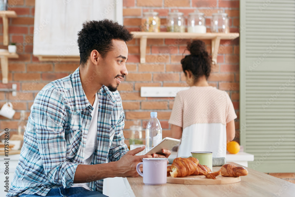 Portrait of mixed race male wears checkered shirt sends text message from his digital tablet computer, uses wireless internet, enjoys sweet croissants and cappucino, sits over kitchen interior Portrait of mixed race male wears checkered shirt sends text message from his digital tablet computer, uses wireless internet, enjoys sweet croissants and cappucino, sits over kitchen interior