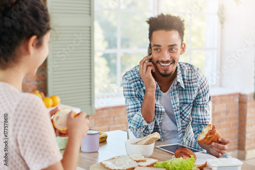 Portrait of joyful man being glad hear old best friend over mobile phone, eats croissant and looks at wife who peels orange. Businessman in casul clothes being at home, arranges meeting with partners photo