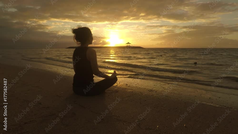 Silhouette of a young woman meditating in lotus pose while sitting on Sanur beach at sunrise time, Bali, Indonesia Silhouette of a young woman meditating in lotus pose while sitting on Sanur beach at sunrise time, Bali, Indonesia